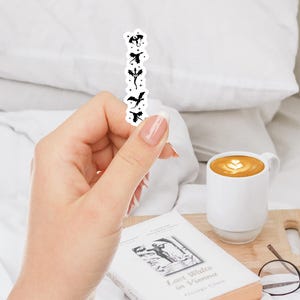 May include: A black and white sticker featuring a vertical row of dragon silhouettes. The sticker is held in a hand, with a white mug of coffee, a book, and glasses in the background.