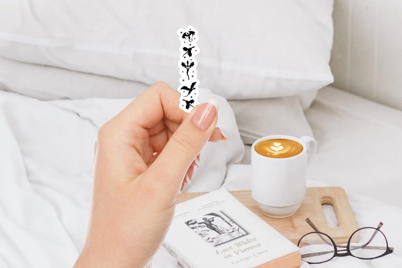 May include: A black and white sticker featuring a vertical row of dragon silhouettes. The sticker is held in a hand, with a white mug of coffee, a book, and glasses in the background.