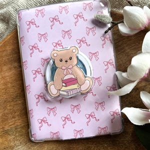 May include: A clear phone case with a pink bow pattern and a teddy bear pop socket. The pop socket features a brown teddy bear with a pink bow, sitting on a stack of books.