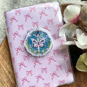 Op de afbeelding: Een rechthoekige tablethoes met een lichtroze achtergrond en een herhalend patroon van kleine roze strikjes. In het midden is een ronde telefoonhouder bevestigd met een afbeelding van een zwaard, een vlinder met roze vleugels en een maansikkel.