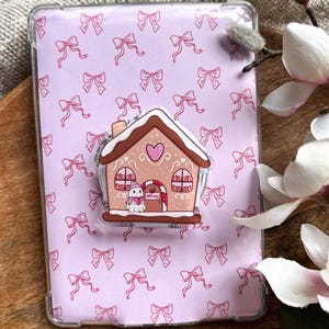 Puede incluir: Una funda transparente para tableta con un patrón de lazos rosas. Un soporte para teléfono con forma de casa de jengibre está adherido a la funda. La casa de jengibre tiene un corazón, ventanas y un pequeño muñeco de nieve.