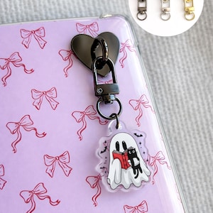 Può includere: Un ciondolo a forma di cuore nero per telefono con chiusura, attaccato a una custodia trasparente con un motivo a fiocco rosa. Un fantasma e un gatto nero leggono un libro sul ciondolo. Tre ciondoli con cerniera sono in alto a destra.