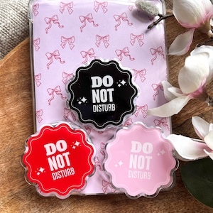 Könnte beinhalten: Eine transparente Hülle mit einem rosa Schleifenmuster, die drei "Do Not Disturb"-Acrylstifte enthält. Die Stifte sind schwarz, rot und rosa, jeweils mit weißem Text und Sternakzenten. Die Hülle liegt auf einer Holzoberfläche.