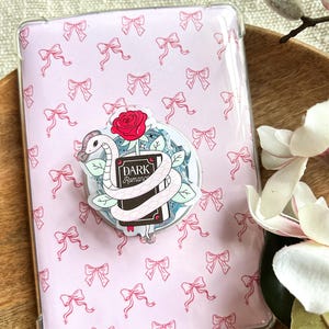 May include: A clear phone case with a pink bow pattern. A circular pop socket features a white snake wrapped around a book with the words "Dark Romance" and a red rose.