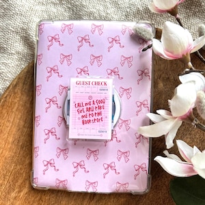 Puede incluir: Una funda de tableta rosa claro con un patrón repetido de lazos rojos. Una pegatina de "Guest Check" está adjunta, con el texto "CALL ME A GOOD GIRL AND TAKE ME TO THE BOOKSTORE". La funda está sobre una superficie de madera, con flores blancas y rosas.