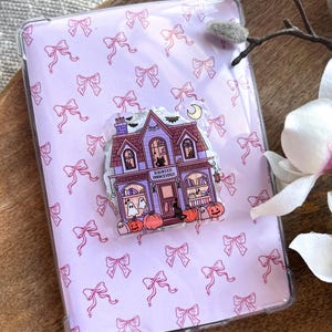 May include: A rectangular tablet case with a light pink background featuring a repeating pattern of small pink bows. Centered on the case is an acrylic charm depicting a purple haunted bookstore with bats, ghosts, pumpkins, and the text Haunted Bookstore.