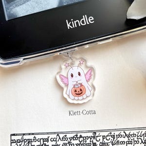 Puede incluir: Un charm de acrílico transparente con un fantasma de dibujos animados con alas rosas y una calabaza. El charm está colgado cerca de un lector Kindle. La palabra "kindle" está impresa en el lector. El texto "Klett-Cotta" está impreso debajo.