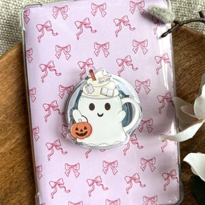 Puede incluir: Una funda transparente para teléfono con un fondo lila claro cubierto de lazos rosas. Un pop socket circular presenta un fantasma de dibujos animados sosteniendo una calabaza y una taza de café con el texto "Pumpkin Spice Latte".