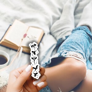 May include: A white sticker featuring black dragon silhouettes in various poses. The sticker is held in a hand with white nail polish, with a book, coffee cup, and denim jeans in the background.