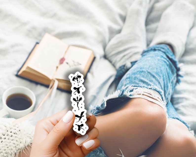 May include: A white sticker featuring black dragon silhouettes in various poses. The sticker is held in a hand with white nail polish, with a book, coffee cup, and denim jeans in the background.