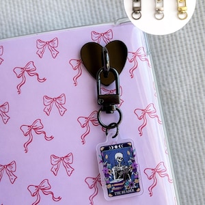 May include: A black heart-shaped clasp with a key ring holding a tarot card-themed keychain. The keychain features a skeleton illustration and the text "THE READER". Three zipper pull charms in black, silver, and gold are also shown.