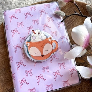 May include: A clear phone case with a light purple bow pattern. A circular pop socket with a cartoon fox in a coffee cup design is attached. The fox is orange and the cup has whipped cream and a straw.