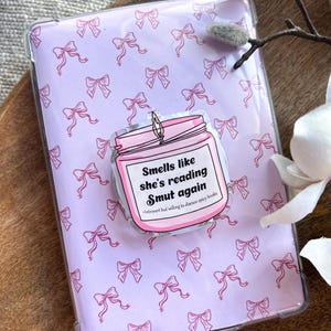 May include: Clear tablet case with a light purple background covered in pink bows. A pink jar sticker with the text "Smells like she's reading Smut again" is centered on the case. The sticker also says "Introvert but willing to discuss spicy books."