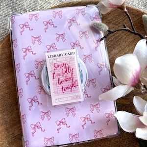 Puede incluir: Una funda transparente para teléfono con un patrón de lazos morados claros, que sostiene una tarjeta de biblioteca rosa que dice "Sorry, I'm fully booked tonight!". La funda está sobre una bandeja de madera con flores de magnolia.
