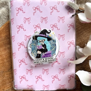 Pode incluir: Uma capa de tablet rosa claro com um padrão de laços rosa. Um suporte pop circular apresenta uma bruxa de desenho animado com cabelo azul, óculos e um chapéu de bruxa, lendo um livro. O texto diz "JUST A WITCH WHO LOVES BOOKS."