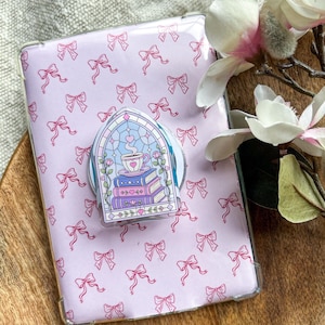 Op de afbeelding: Een rechthoekige tablethoes met een lichtpaarse achtergrond en een herhalend patroon van roze strikken. In het midden is een telefoonhouder bevestigd met een glas-in-loodontwerp van een theekopje op een stapel boeken met bloemen.