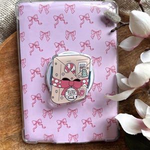 Puede incluir: Una funda transparente para teléfono con un fondo lila claro cubierto de lazos rosas. Un agarre emergente para teléfono presenta un diseño rosa y blanco con un lazo, un bastón de caramelo y el texto "Blind Date with a Book".