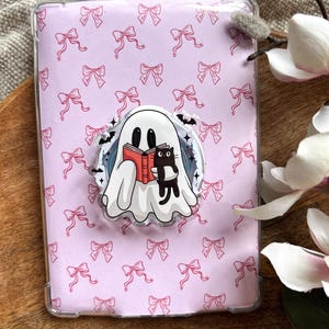 Peut inclure: Une coque de tablette rose clair avec un motif répétitif de nœuds roses. Un autocollant circulaire représente un fantôme de dessin animé lisant un livre rouge avec un chat noir. La coque est dans une housse de protection transparente.