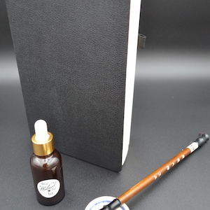 Könnte beinhalten: Ein schwarzes, ledergebundenes Notizbuch mit weißem Buchschnitt steht neben einer Flasche brauner Flüssigkeit mit weißem Etikett. Ein Kalligraphiepinsel liegt auf einer kleinen Keramikschale. Der Pinsel hat einen Holzgriff und eine weiße Spitze.