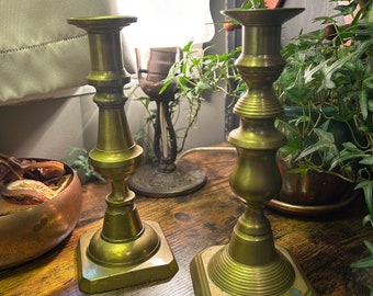 May include: Two antique brass candle holders with a fluted design and square bases. The candle holders are on a wooden table.