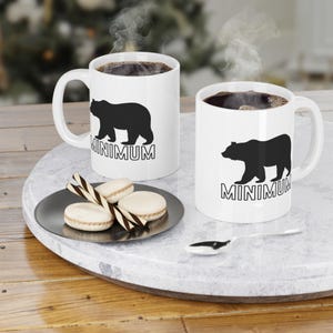 Puede incluir: Dos tazas de cerámica blanca con siluetas de osos negros y el texto "MINIMUM" impreso en ellas. Las tazas están llenas de café y sale vapor de la parte superior de cada taza. Las tazas están sobre una superficie de mármol blanco con un pequeño plato de galletas.