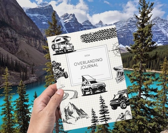 May include: A hand holding a journal with the text "OVERLANDING JOURNAL 2026". The journal features illustrations of off-road vehicles, mountains, trees, and a compass. The background shows a scenic view of mountains, a lake, and trees.