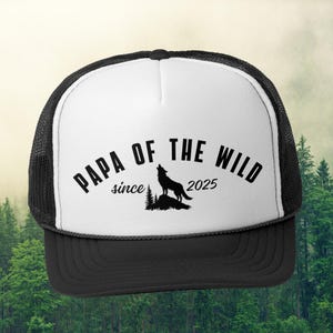 Könnte beinhalten: Schwarz-weiße Trucker-Kappe mit schwarzem Netzrücken. Die Vorderseite der Kappe hat einen weißen Hintergrund mit dem Text "Papa of the Wild since 2025" in Schwarz. Eine Silhouette eines heulenden Wolfes befindet sich in der Mitte des Textes.