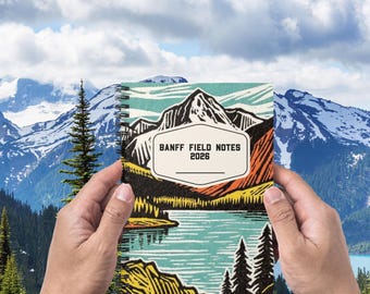 May include: A spiral-bound notebook with a mountain and lake landscape design. The cover features the text "BANFF FIELD NOTES 2026" in a white rectangular box. The notebook is held against a backdrop of mountains and a cloudy sky.