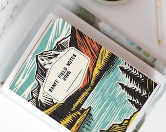 May include: A notebook with a colorful mountain landscape design. The cover features a stylized illustration in shades of orange, yellow, blue, and black. The text "BANFF FIELD NOTES 2026" is printed on a white label. A pen and a cup are visible in the background.