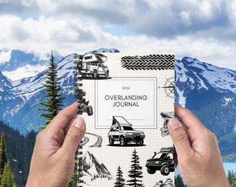 May include: A spiral-bound journal with the text "OVERLANDING JOURNAL 2026" on the cover. The cover features illustrations of off-road vehicles, mountains, trees, and a compass. The background shows a mountain range and a cloudy sky.