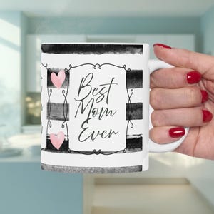 May include: A white ceramic mug featuring a black and white horizontal striped pattern. The center displays a decorative black frame containing the text Best Mom Ever in a cursive script, accented by two small pink hearts on the left side.