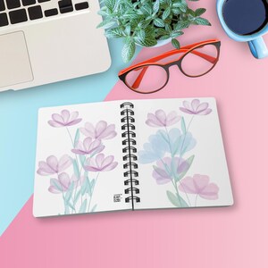 Può includere: Un taccuino a spirale con un'illustrazione floreale in stile acquerello con petali viola chiaro e steli verde pallido su entrambe le pagine. Il taccuino include un piccolo codice a barre nero vicino alla rilegatura a spirale centrale.