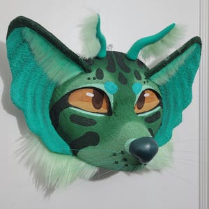 May include: A green and teal furry mask with large ears and horns. The mask has orange eyes, a dark green nose, and long whiskers. The mask is decorated with black spots and light green fur around the ears and chin.
