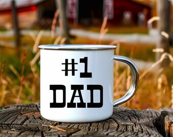 May include: White enamel camping mug with a black #1 DAD design. The mug is sitting on a wooden surface with a red barn in the background.