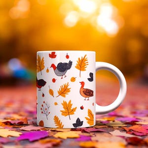 May include: White ceramic mug featuring a fall-themed design with illustrations of birds, leaves, and berries in orange, red, and black. The mug has a white handle and is set against a blurred background of autumn leaves.