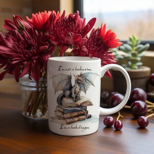 May include: White ceramic mug with a blue and gray dragon sitting on a stack of books. The mug has the text "I'm not a bookworm, I'm a bookdragon." printed on it.