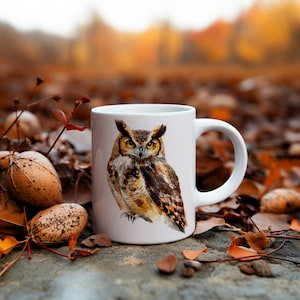 May include: White ceramic mug with a brown and yellow owl illustration. The owl is looking directly at the viewer. The mug is sitting on a bed of brown leaves.
