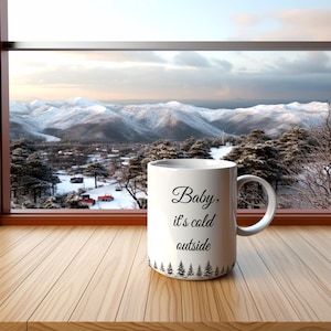 May include: White ceramic mug with black text that reads "Baby, it's cold outside" and a black silhouette of pine trees at the bottom. The mug is sitting on a wooden surface in front of a window with a view of snow-covered mountains.