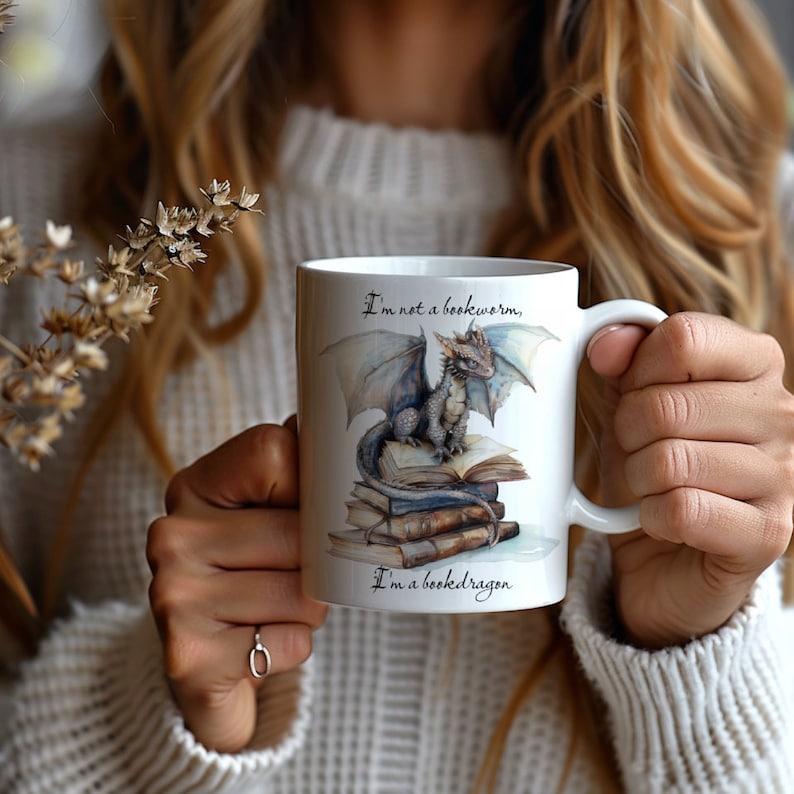 May include: White ceramic mug with a blue and gray dragon illustration sitting on a stack of books. The mug has the text "I'm not a bookworm, I'm a book dragon."