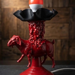 May include: A red table lamp with a unique design. The lamp features a red goat figurine, a textured red column, and a black vinyl record base supporting a white and orange light bulb. The lamp is plugged in with a white cord.