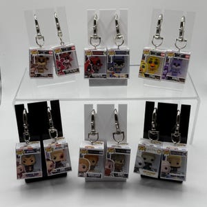 May include: Silver-tone earrings featuring miniature Funko Pop figures in clear display boxes. The collection includes characters like Freddy, Deadpool, and characters from Avatar and Harry Potter. The earrings are displayed on a white surface.
