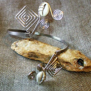May include: Silver-toned wire arm cuff featuring spiral and square designs, accented with white seashells. The cuff is displayed on a piece of driftwood, set against a textured, neutral background.