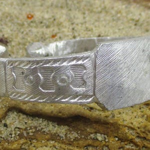 Op de afbeelding: Een zilverkleurige ring met een rechthoekige voorkant en gegraveerde geometrische ontwerpen. De ring heeft een getextureerd oppervlak en is geplaatst tegen een zanderige achtergrond. De band van de ring heeft een decoratief patroon.