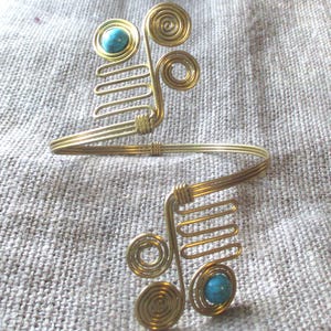 May include: Gold-toned arm cuff with a unique design. The cuff features spiral and linear wire elements, with a turquoise-colored stone accent at each end. The piece is set against a neutral fabric background.