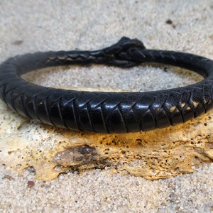 Puede incluir: Un brazalete de cuero negro con un diseño trenzado. El brazalete está atado con un nudo en el extremo.