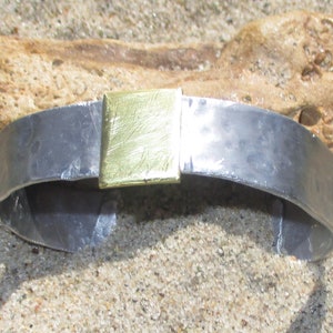 May include: A silver bracelet with a gold rectangular accent. The bracelet is curved and has a smooth, polished finish.