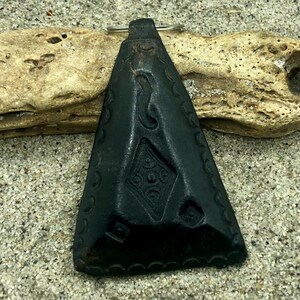 Może przedstawiać: Czarny skórzany wisiorek z misternymi geometrycznymi wzorami i srebrną pętlą. Wisiorek ma kształt trójkąta i teksturę.