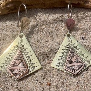 May include: A pair of dangle earrings featuring a gold-toned triangular metal base with a smaller, copper-toned triangular inlay. The surface of each earring is decorated with etched black line patterns, and they hang from silver-toned metal hooks.