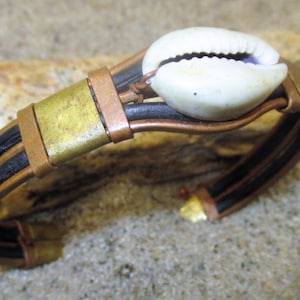 May include: A handcrafted bracelet featuring a white seashell centerpiece. The bracelet has black leather bands, accented with copper and brass metal details. The design evokes a natural, bohemian aesthetic.