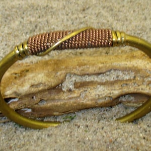 Puede incluir: Un brazalete dorado con un acento de alambre de cobre. El alambre está envuelto alrededor del brazalete y crea un diseño decorativo.